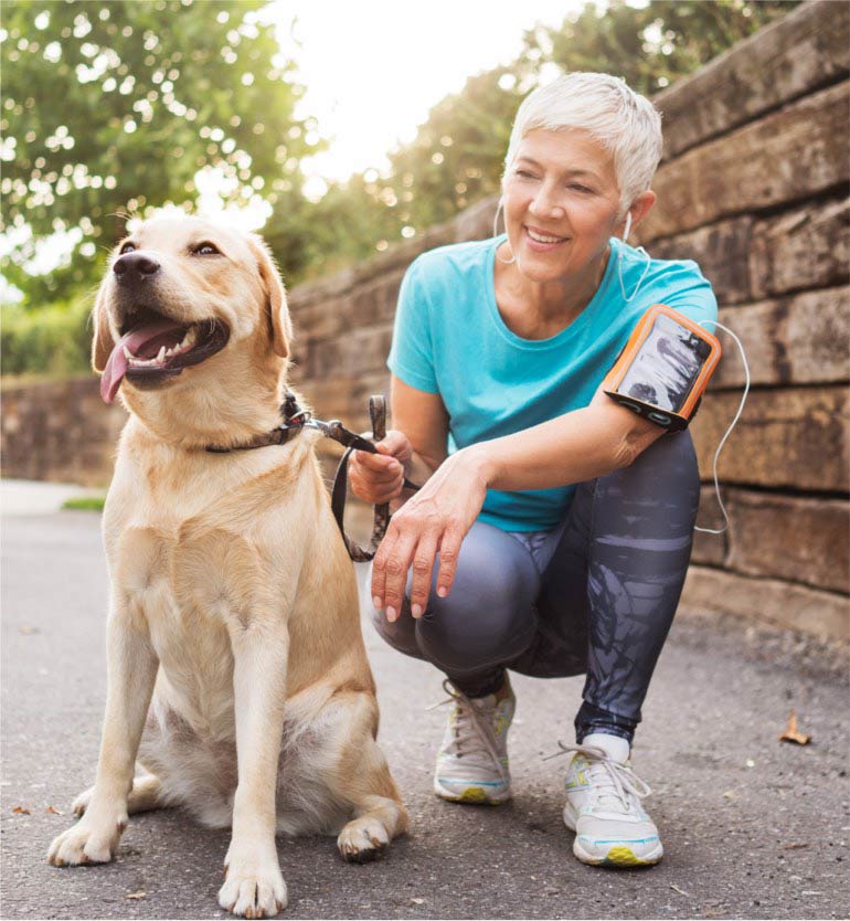 A senior woman in sportswear kneeling beside a happy Labrador on a leash during a walk.