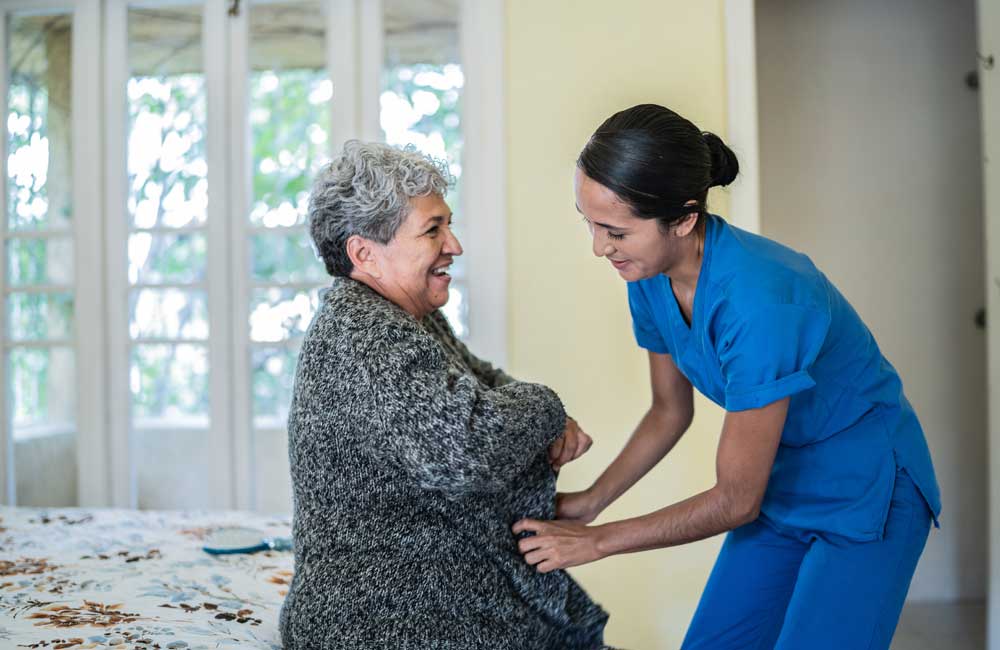 Caregiver assisting an elderly woman with her sweater while both are smiling warmly at each other.
