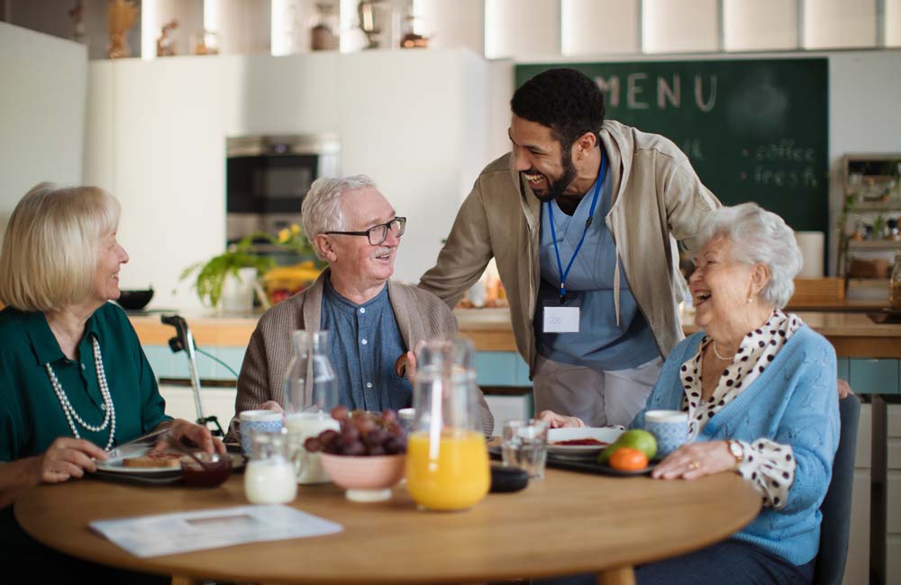 A caregiver engages with three elderly adults at a dining table in a memory care facility.