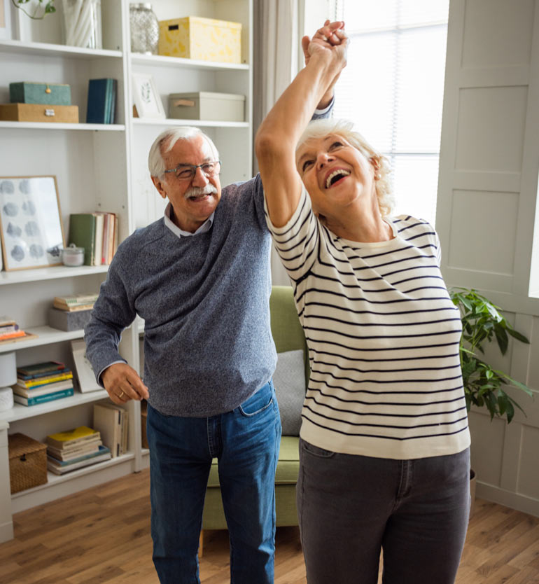 Elderly couple smiling and dancing together in a cozy, bright living room with bookshelves and plants.