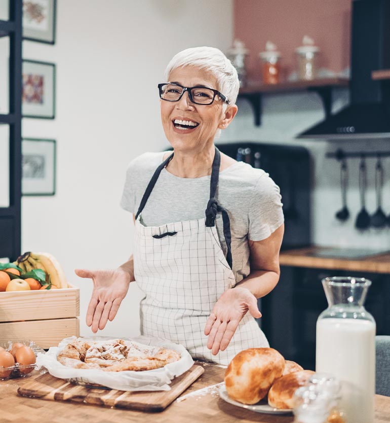 Elderly woman in a checkered apron smiling in a kitchen, displaying freshly baked pastry and bread.