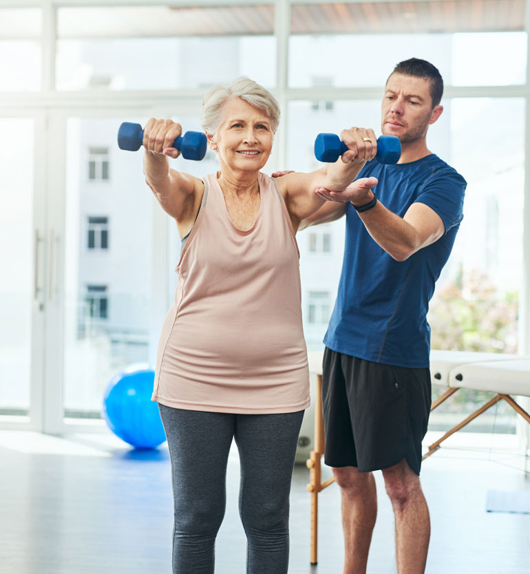 Elderly woman lifting dumbbells with assistance from a trainer in a bright gym setting.