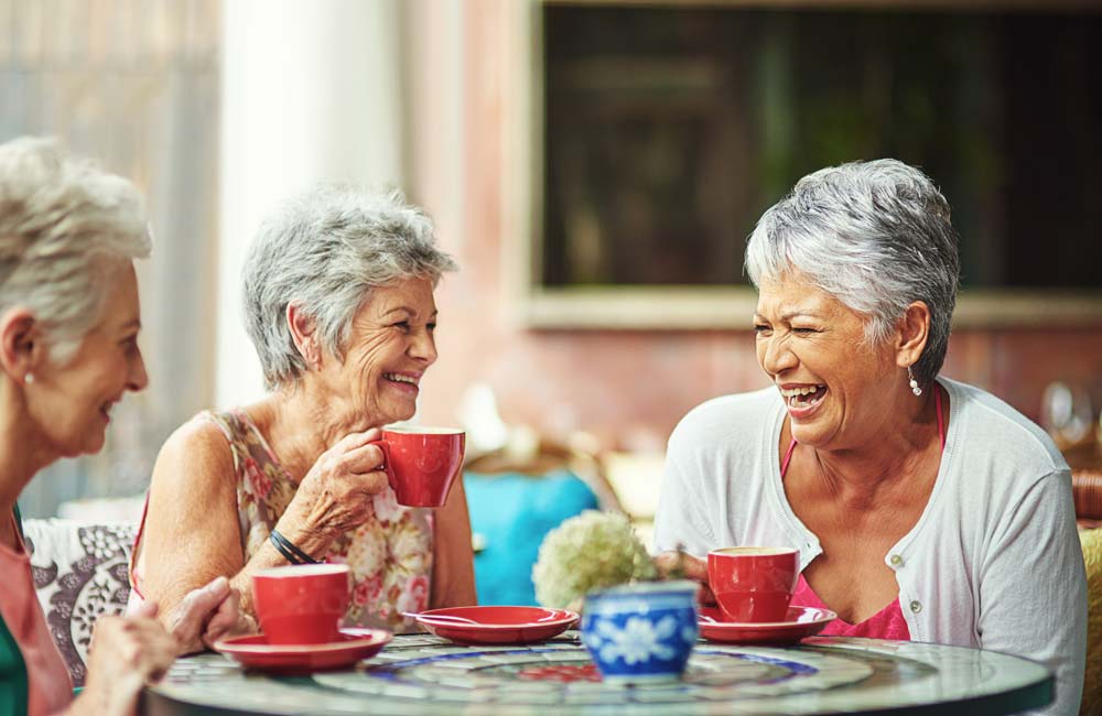 Three elderly women enjoying coffee and laughing together at an outdoor cafe.