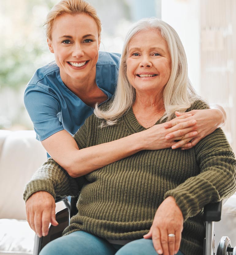 Smiling healthcare worker embracing and supporting a happy senior woman in a wheelchair.