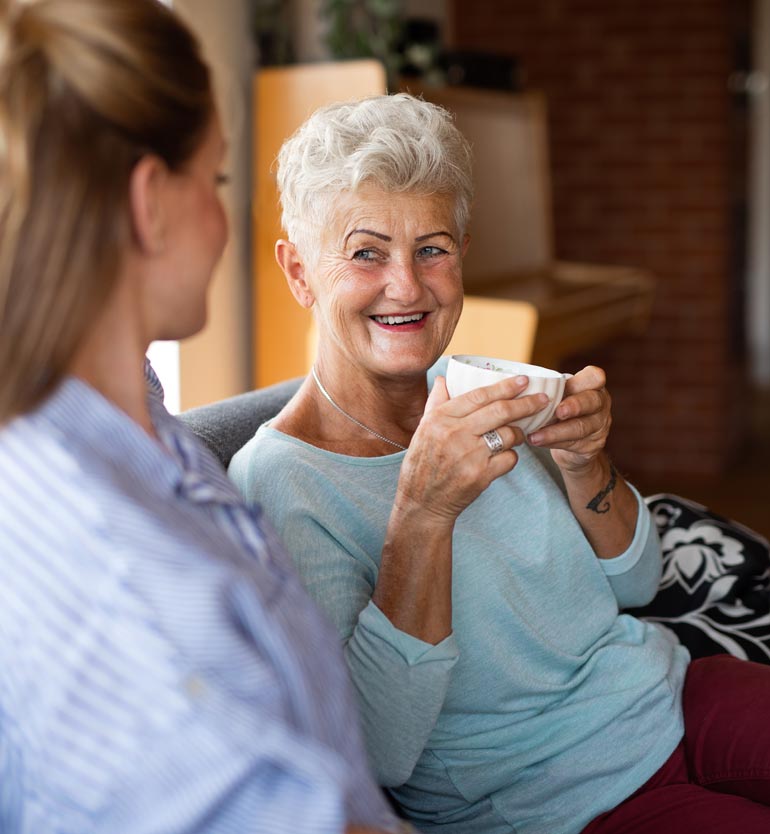 Older woman smiling while holding a cup of tea, conversing with a younger woman in a relaxed setting.