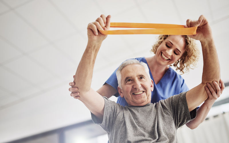 Senior man exercising with resistance band assisted by a smiling caregiver in a bright room.
