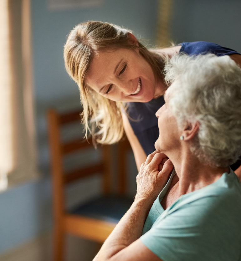 Caregiver smiling at elderly woman in a cozy room, fostering a compassionate environment.