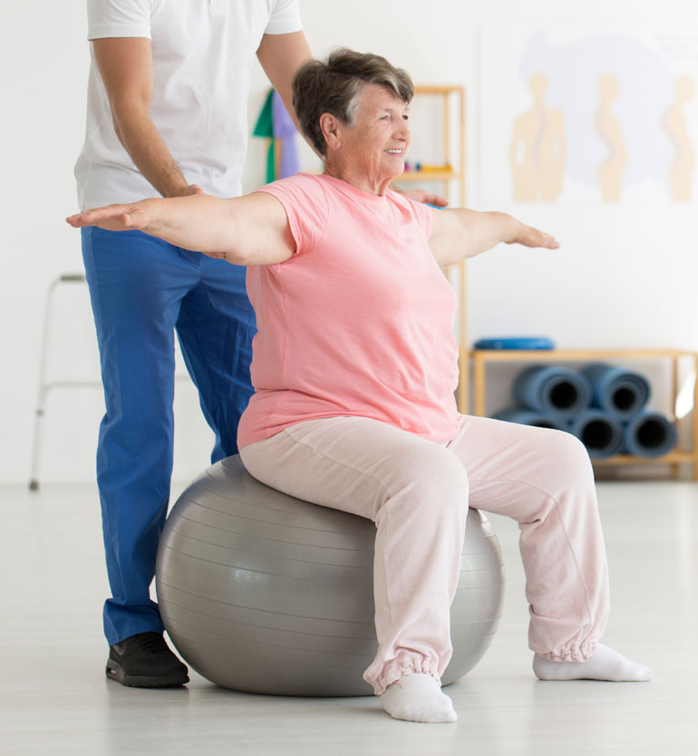 A senior woman doing arm exercises on a stability ball during a therapy session with a therapists support.
