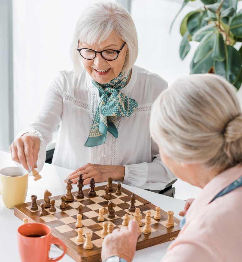 Two senior women, one wearing glasses, enjoy playing chess together at a brightly lit table.
