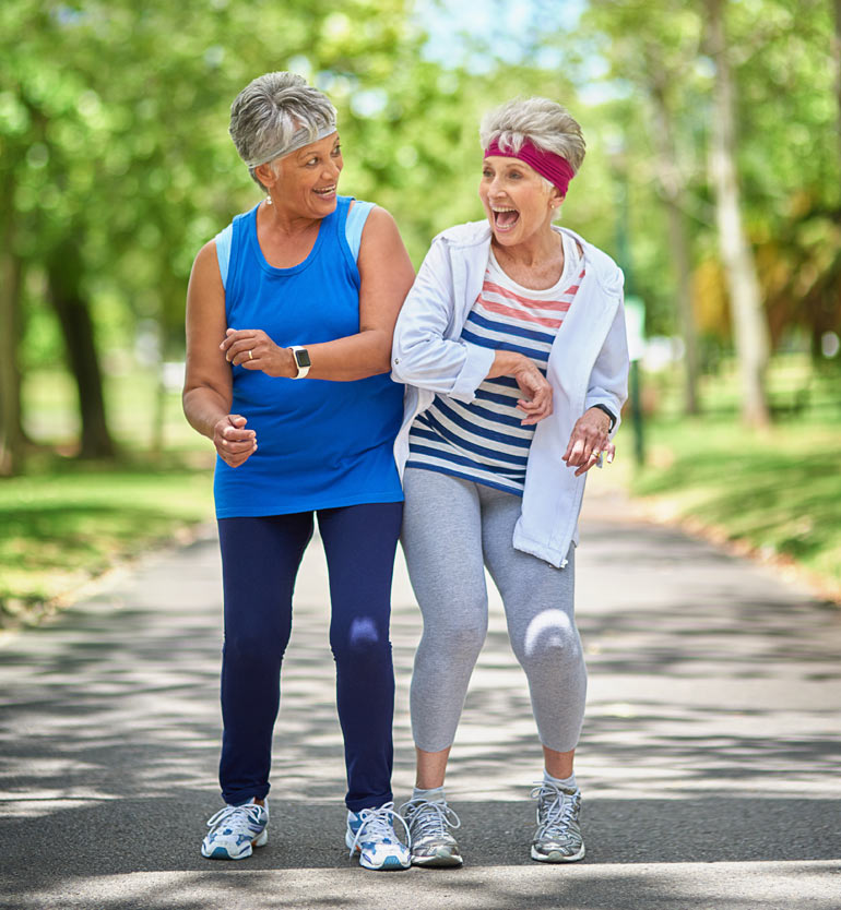 Two senior women laughing and power-walking together on a park trail in vibrant athletic wear.