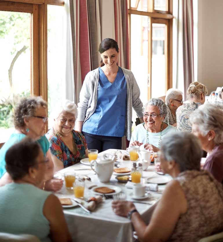 A group of seniors dining together, supervised by a friendly caregiver at an assisted living facility.